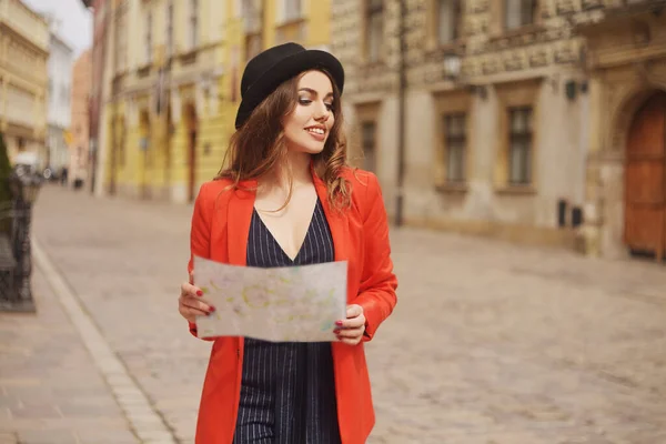 快乐的女人带着地图在度假 旅行者漫步在城市 阳光灿烂的日子 后视镜一个戴着帽子 穿着红色夹克的年轻女游客 站在城市街道上的模特儿正在地图上寻找一条路 — 图库照片