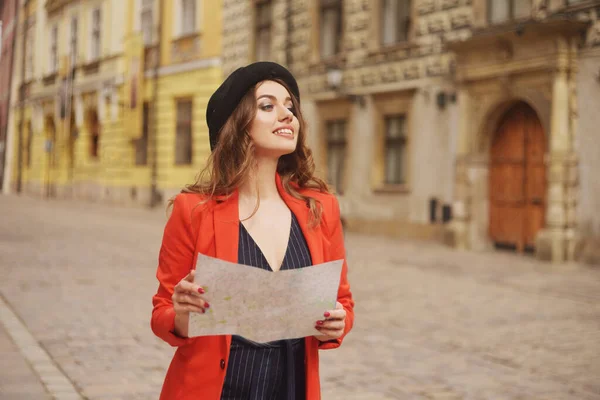 Happy woman on vacation with map, traveler walks the city. Sunny day. Back view. A young tourist woman in a hat, red jacket. Model girl standing on a city street and is looking for a way on the map.