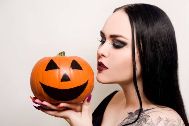 Güzel bir esmer, elinde bir balkabağı tutuyor ve ona bakıyor. Cadılar Bayramı makyajlı, siyah rujlu bir kadın. Beyaz arkaplanda model.