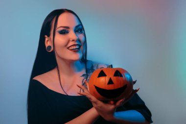 Gülümse Cadılar Bayramı cadısı kameraya bakar ve elinde bir balkabağı tutar. Neonlu falcı. Dövmeli ve piercingli esmer kadınlar kameraya bakıyor..