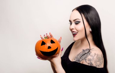 Güzel bir esmer, elinde bir balkabağı tutuyor ve gülümsüyor. Cadılar Bayramı kostümlü partisi. Kirpiklerin için örümcek ağı, cadı. Beyaz fotoğraftaki kadın.