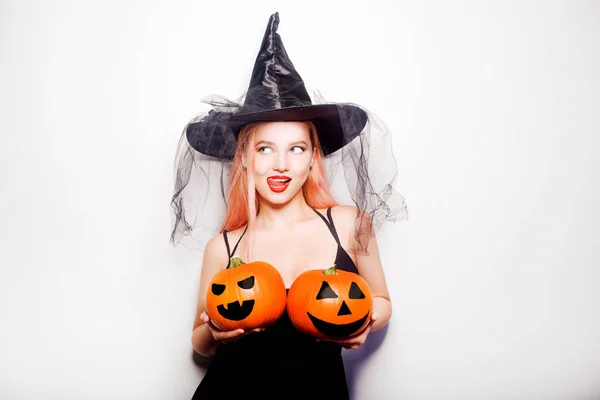 Woman in witch costume, keep pumpkin covering her breasts. Funny photo, invitation to Halloween party. The model looks to the side at an empty space