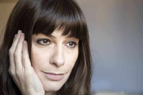 35 years old woman close-up portrait. Caucasian with brown long hair and bangs. Smiling, serene expression, looking down with an hand on face.