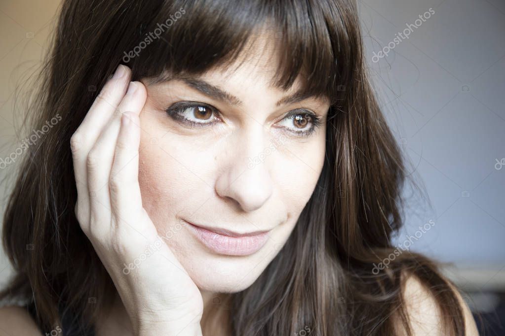 Young woman close-up portrait. Caucasian with brown long hair and bangs. Serene expression, looking down with one hand on face.