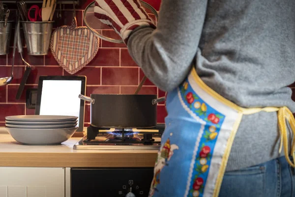 Kırmızı klasik mutfak, gri bir tencerede yemek tarifi tablet okunurken önlük giyen kadın
