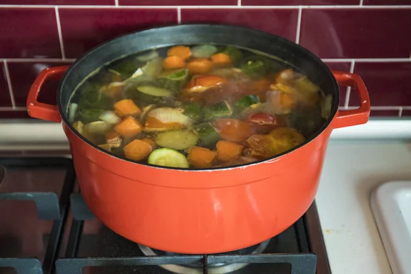 Red pot with boiling vegetable soup on the stove in burgundy kitchen.