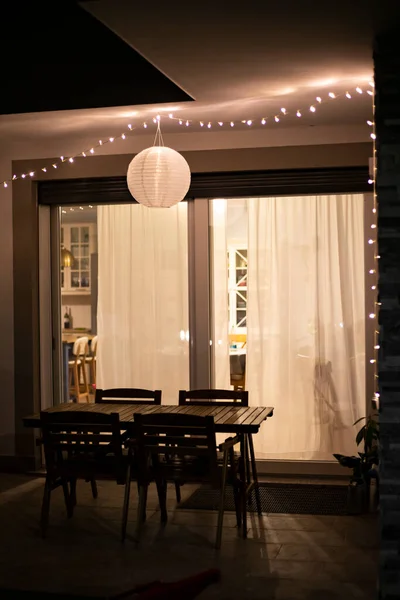 Inspiration home decor idea, with string lights and a globe solar lantern hanging on the ceiling in the front porch, over outdoor furniture, warming the backyard or the garden in summer nights. Vertical.