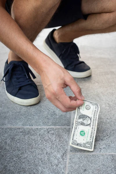 İsimsiz bir adam, sıradan giyinmiş, mavi ayakkabılı, yerden bir dolarlık banknot alıyor. Dikey çekim.