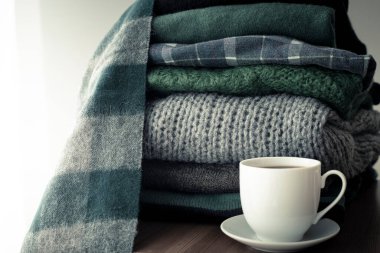 Yığınla yeşil ve gri süveter, yanında sıcak içecek beyaz bardağı, arka planında fotokopi alanı, rahat, hygge ya da mevsimlik gardırop değiştiren duvar kağıdı. Yeşil filtre, tonlu resim.