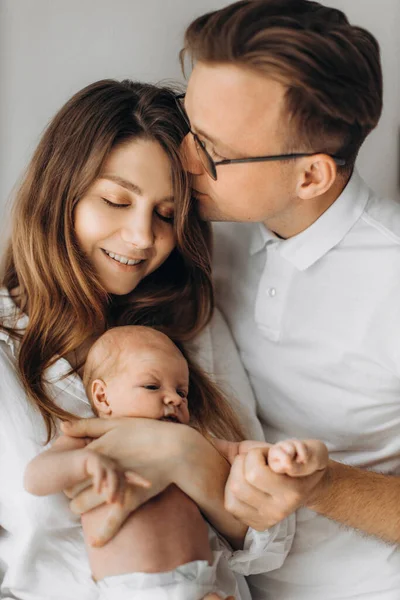 Счастливые родители с новорожденной девочкой, любящая мать держит маленькую дочь на руках, заботливый муж целует великолепную жену, наслаждаются счастливыми моментами, концепция молодой семьи — стоковое фото