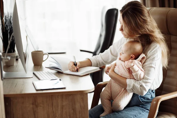 Komoly fiatal anya ül az asztalnál, tartsa a karját kislánya, gyönyörű hölgy jegyzetel, tervezés, leírja ötletek, üzletasszony dolgozik az otthoni online — Stock Fotó