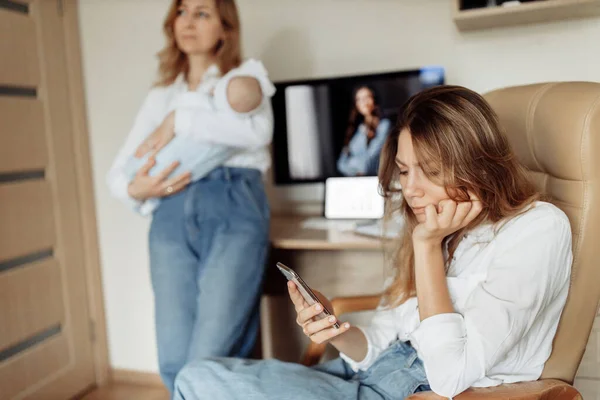 深刻な女性は椅子に座って、笑顔で、スマートフォンを使用して、良いニュースを読んで、若い母親は自宅でオンラインで働いています。 — ストック写真