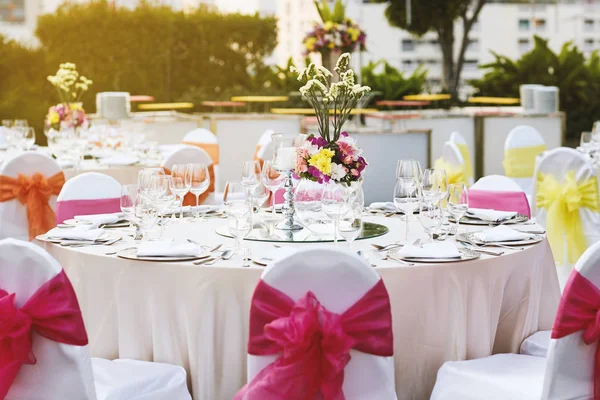 Warm sunset light with wedding reception dinner table setting with empty glasses of water, wine and dinnerwear with flower decoration, and white fabric cover chairs with pink sash