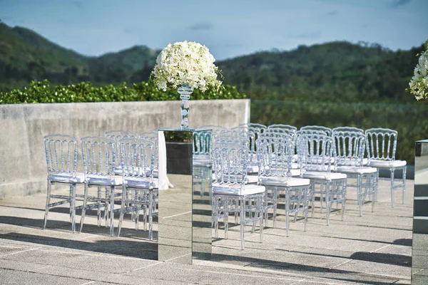 Beach wedding venue setup, ghost chairs setting with roses and white flower on the mirror box vase decoration with mountain background