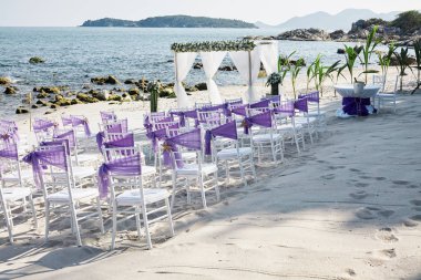 Beach wedding venue settings on the white sand at seaside with white chiavari chairs decorating with violet organza sash, minimal flower decoration, panoramic blue sea on sunny day clipart