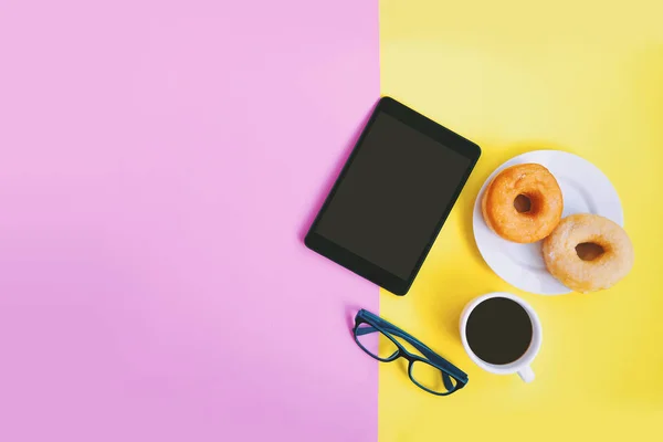 Modern home office workspace, Black coffee cup and donuts, eye glasses and tablet on pink and yellow pastel background with copy space