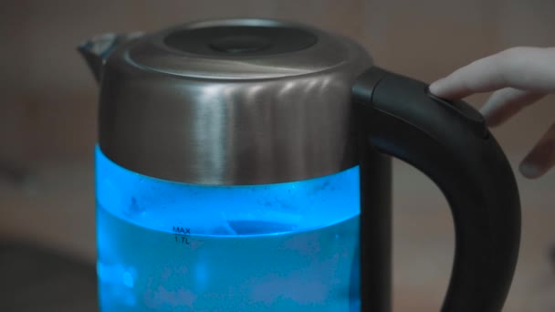 Czajnik elektryczny ze szkła z światła czyraki. Niebieski Woda wrze w zbliżeniu czajnik. Ręka zawiera piękny czajnik elektryczny. wideo 4k — Wideo stockowe