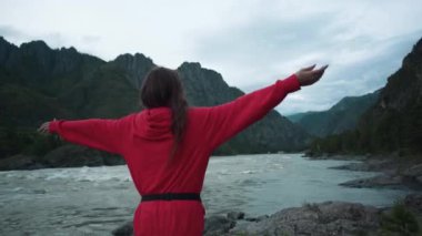Güzel bir kız nehrin kıyısında durur ve kollarını uzatır. Dağlarda bir kadın kayalıklarda duruyor. Altai, Rusya, 4k video.