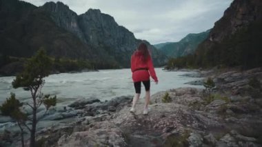 Güzel bir kız nehrin kıyısında durur ve kollarını uzatır. Dağlarda bir kadın kayalıklarda duruyor. Altai, Rusya, 4k video.