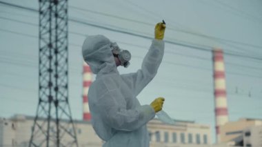 Bilim adamı bir solunum cihazı ve koruyucu bir bornoz giydi. Bir öğrenci fabrikanın arka planındaki ekolojiyi kontrol ediyor. Zararlı kimyasallar havayı kirletir. Novosibirsk. Coronavirus salgınında, COVID-19.