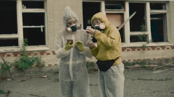 Zwei Wissenschaftler legen eine Atemschutzmaske an und forschen. Die Umwelt ist in Gefahr. Coronavirus in einer Pandemie, COVID-19. Zwei Mädchen in Schutzanzügen vor Strahlung. Geben Sie die Daten ins Telefon ein. — Stockvideo