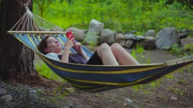 Tatilde bir hamakta yatan rahat genç güzel kız. Bir kadın telefonda fotoğrafları üzerinden kaydırır, bir SmartWatch 'da fotoğrafları. 4k video.