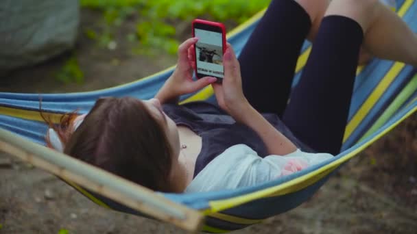 放松的年轻美丽的女孩躺在吊床上度假 一个女人在手机上滚动浏览照片 在智能手表上拍摄自己 — 图库视频影像