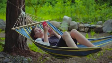 Tatilde bir hamakta yatan rahat genç güzel kız. Bir kadın telefonda fotoğrafları üzerinden kaydırır, bir SmartWatch 'da fotoğrafları. 4k video.