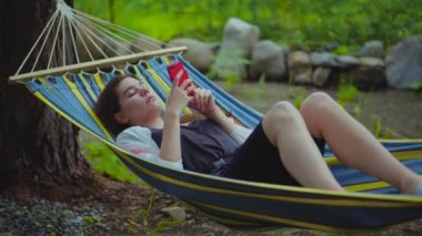 Tatilde bir hamakta yatan rahat genç güzel kız. Bir kadın telefonda fotoğrafları üzerinden kaydırır, bir SmartWatch 'da fotoğrafları. 4k video.