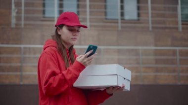 Kırmızı üniformalı bir pizza cıvıltIsı iki kutu pizza getirmiş. Mutlu kız telefonla sipariş netleştirmek için müşteriye geri çağırır. Bir tuğla evin arka planına karşı.