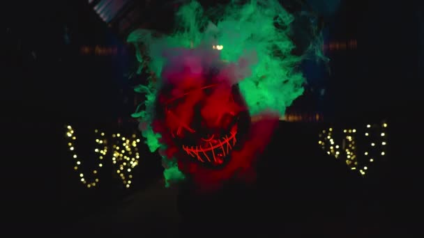 El hombre de la máscara de neón sopla humo de la purga. Humo grueso verde y rojo. Traje brillante para halloween. — Vídeo de stock