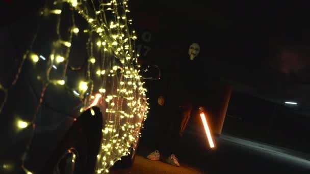 Człowiek w przerażającej białej masce trzyma się lampy neonowej. Mordercy nocą na parkingu na tle samochodu z girlandą. Samochód jest urządzony na Halloween. — Wideo stockowe