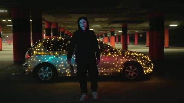 Mannen med läskig vit mask håller i ett baseballträ. Mördare på natten på parkeringen mot bakgrund av en bil med en krans. Blodiga halloween karneval kostym. — Stockvideo