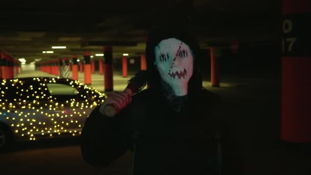 Un hombre con una máscara blanca aterradora sostiene un bate de béisbol. Asesinos por la noche en el estacionamiento contra el telón de fondo de un coche con una guirnalda. Disfraz de carnaval de Halloween sangriento. — Vídeo de stock