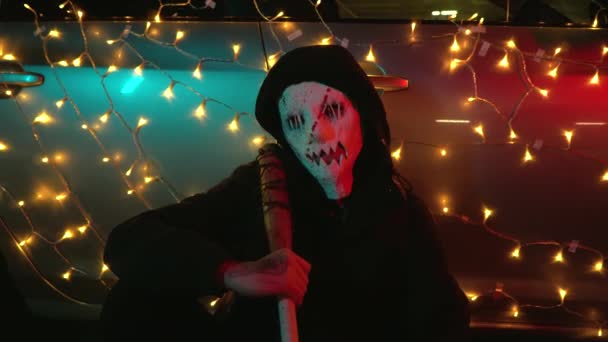 A man in a bloody mask sits on the pavement. The killer is ready for the Purge. A baseball bat wrapped in barbed wire for Halloween. The dangerous car is decorated with a garland. — Stock Video