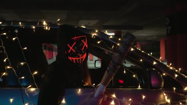 Mężczyzna w neonowej masce wygląda przez okno samochodu na parkingu w nocy. Zabójca trzyma kij baseballowy. Przerażający kostium z czystki na Halloween. — Wideo stockowe