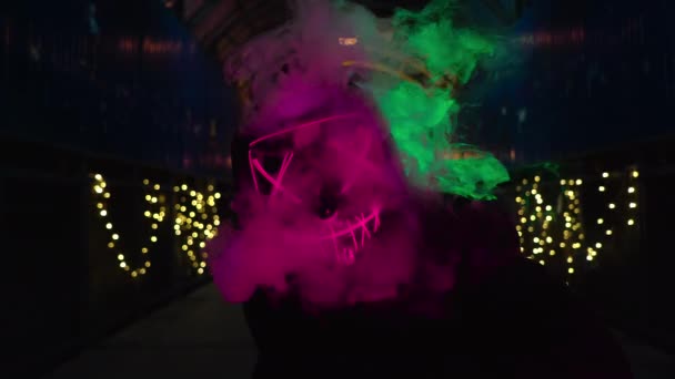 Człowiek w neonowej masce dmucha dym. Gruby zielony i różowy dym. Przerażający kostium zabójcy Halloween Purge. — Wideo stockowe