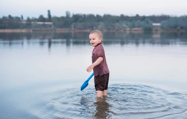 The kid in clothes stands in the lake . A toy of children for beach rest in hands of the beautiful boy