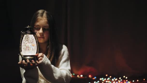 かわいい女の子は彼女の腕でクリスマス ランタンを押しながら暗い部屋で床に座って 雪とクリスマス グローブ ランタンのライトを見てください — ストック動画