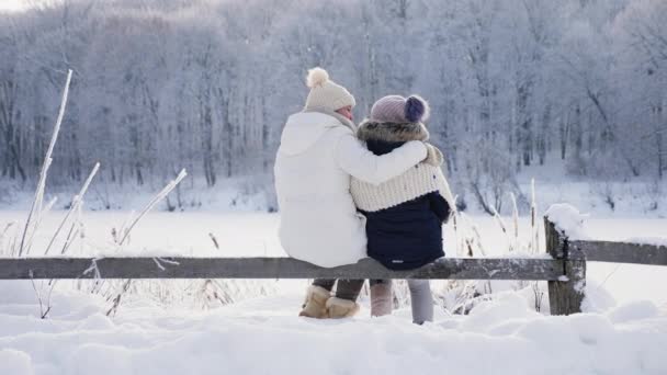 后面的风景妈妈和女儿穿着暖和的衣服 拥抱在一起欣赏山区的冬日风景 坐在被雪覆盖的木栅栏上 — 图库视频影像