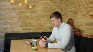 Yan görünümü çekici adam kullanım telefon bir kafede oturuyor. Yakışıklı adam ile smartphone bir tuğla duvara barda oturan