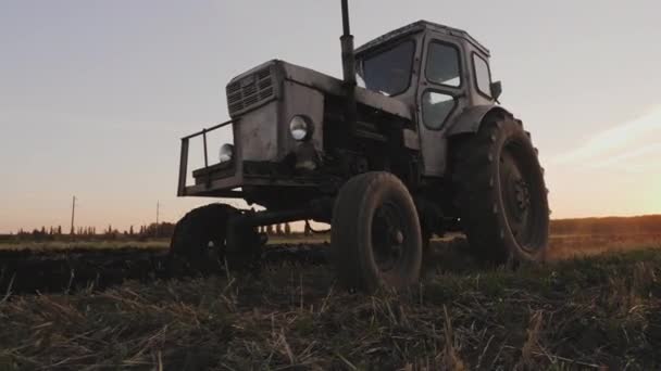 Oude grijze trekker met een harrow aan het werk op een veld bij zonsondergang — Stockvideo