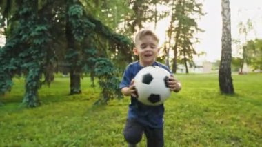 Sevimli küçük bir çocuk bir futbol topu tutarak, uzak yeşil çim üzerinde çalışan ve mutlu gülümseyen. Çekim yavaş hareket çekim