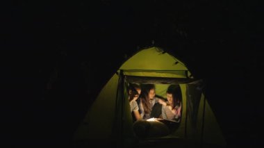 Geceleri kamp çadırında köpekli bir aile kitap okuyor.