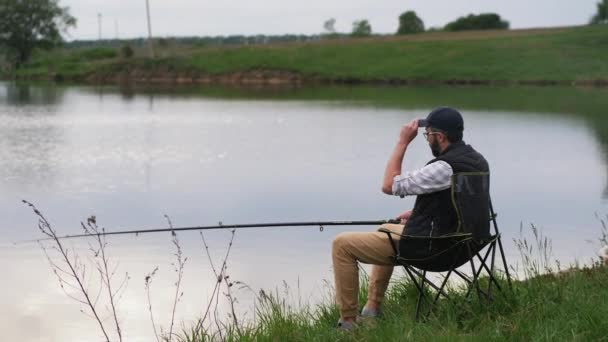 Fischer sitzt mit Angelrute auf einem Stuhl am See — Stockvideo