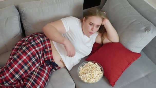 Товста дівчина їсть попкорн, лежачи на дивані — стокове відео