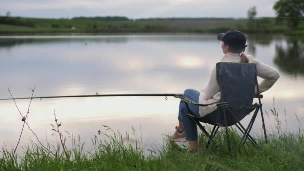 Woman with a fishing rod sitting on a chair by the lake — Stock Video