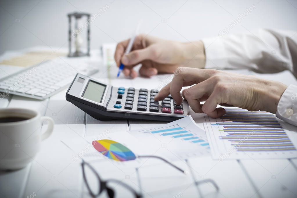 A business person, an accountant, writes a report using a calculator while sitting at a desk in the office.