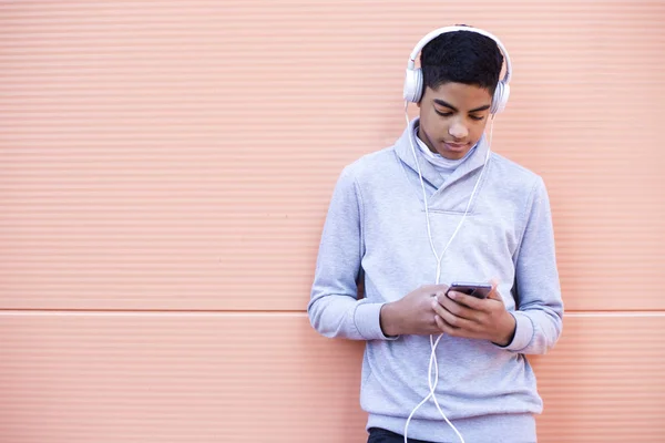 Black young man listens to music on his mobile phone while standing on the street. African American teenager in headphones using a smartphone.