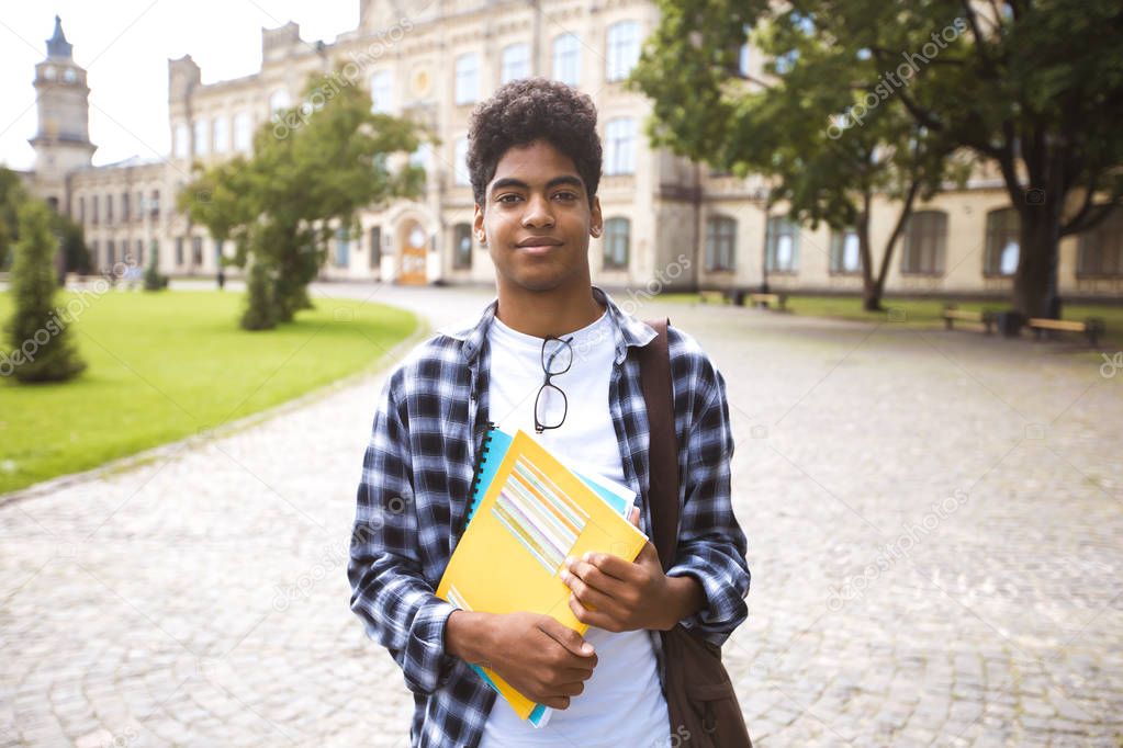 Portrait of a young African American student black man the background of college. Smiling teenage male mixed race keeps books standing near the university.